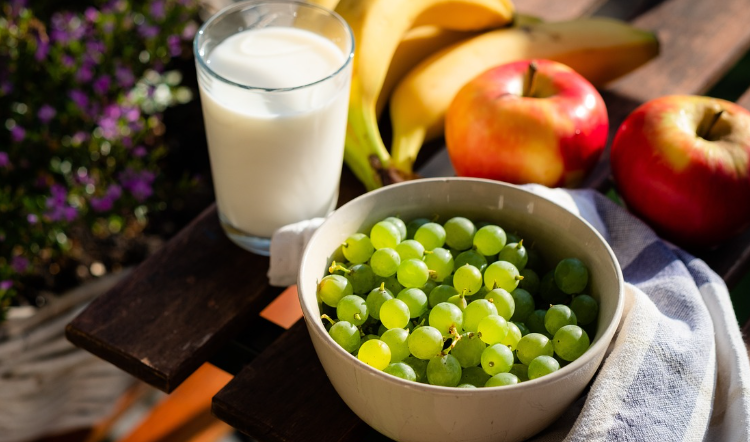 Ciotola di uva verde con bicchiere di latte, mele e banane su tavolo all’aperto. Nutrizione