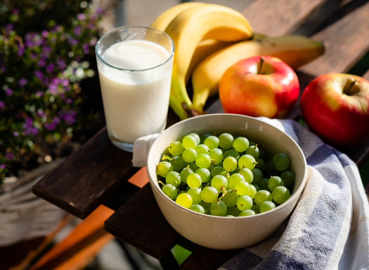 Ciotola di uva verde con bicchiere di latte, mele e banane su tavolo all’aperto. Nutrizione